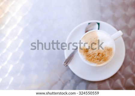 Picture of tasty aroma cappuccino in cup with saucer and spoon. Fresh top of milk in white ceramic on blurred indoor background.