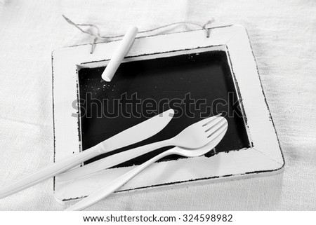 All white place setting concept with white utensils lying on a vintage school slate with white border over textured white textile, high angle view with copyspace on the slate