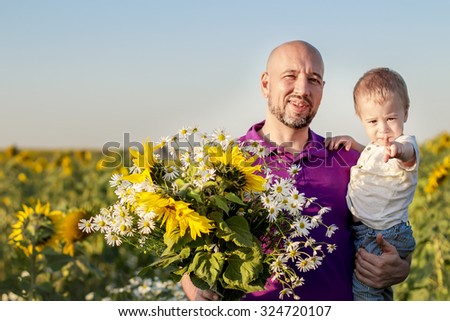 Portrait of bold handsome father holding his little fair hair son and bunch of sunflowers and camomile against sunflower field. Little boy is pointing his finger on you. Image with selective focus