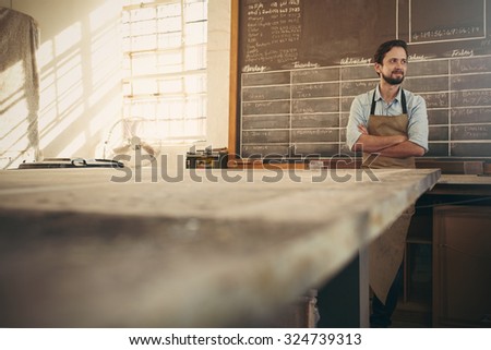 Carpenter craftsman standing with his arms crossed in his workshop with beautiful natural light coming through the window and looking away thoughtfully while smiling confidently