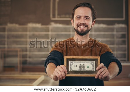 Smiling entrepreneur standing proudly in his workshop and showing you a bank note that has been framed