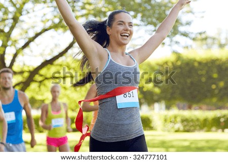 fitness, sport, victory, success and healthy lifestyle concept - happy woman winning race and coming first to finish red ribbon over group of sportsmen running marathon with badge numbers outdoors Royalty-Free Stock Photo #324777101