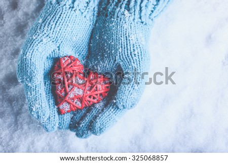Woman hands in light teal knitted mittens are holding a beautiful a entwined vintage romantic red heart in a snow background. Love and St. Valentine concept.