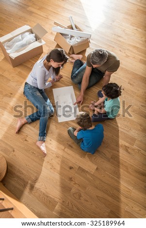 Happy parents are sitting barefoot on the floor of their new house They are looking at the house model with their daughter and son They are surrounded by open cardboard boxes and palette for the paint