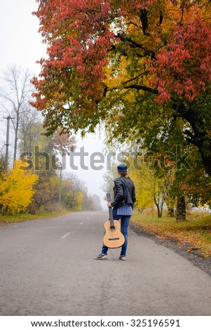 Picture of beautiful young lady in hat with guitar waiting on countryside road. Backview of pretty girl wearing black jacket and jeans on colorful autumn outdoor background.