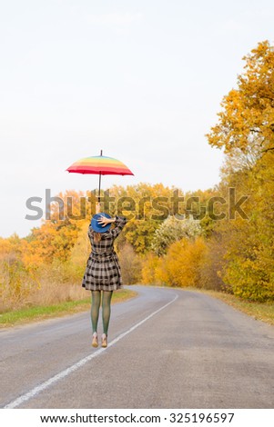 Picture of beautiful young lady with rainbow umbrella jumping on autumn countryside road. Backview of delightful girl in coat on golden trees outdoor background.