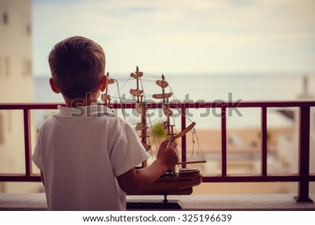 Picture of little kid holding beautiful sailboat model. Backview of dreamy boy on balcony looking at sea on sunset sky outdoor background.