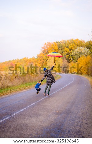 Picture of beautiful young woman with boy holding rainbow umbrella and jumping on autumn countryside road. Backview of happy family having fun on golden trees outdoor background.