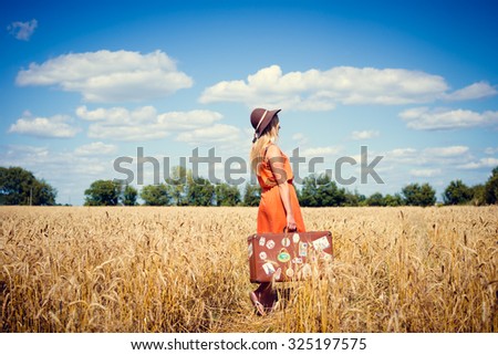 Picture of romantic elegant female in hat holding suitcase in summer wheat field. Charming girl standing on sunny blue sky countryside background.