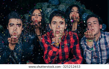 Closeup of young friends group blowing a colorful confetti to the camera in a outdoors party. Friendship and celebrations concept.