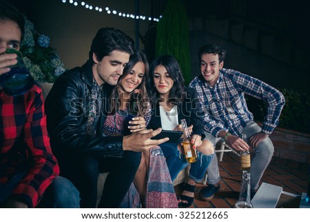 Group of happy young friends drinking and laughing while looking a smartphone picture in a outdoors party. Friendship and celebrations concept.