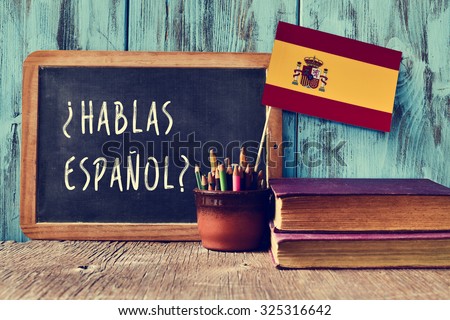a chalkboard with the question hablas espanol? do you speak Spanish? written in Spanish, a pot with pencils and the flag of Spain, on a wooden desk Royalty-Free Stock Photo #325316642