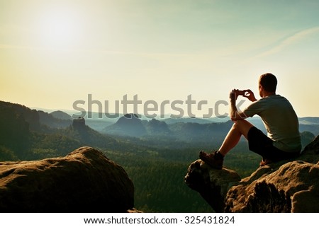 Tourist in grey t-shirt takes photos with smart phone on peak of rock. Dreamy hilly landscape below, spring misty sunrise 