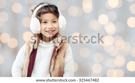 winter, people, christmas, fashion and childhood concept - happy little girl wearing earmuffs and gloves over holidays lights background