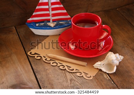 red cup of tea and letter paper next to vintage decorative boat on wooden old table. retro filtered image 
