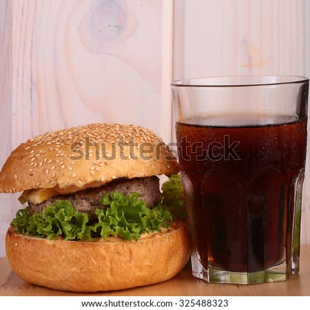 Big tasty appetizing fresh burger of green lettuce bacon slice meat cutlet and white bread bun with sesame seeds on wooden table and cola, square picture