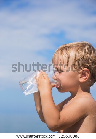 Cute little boy is drinking clean water from a plastic cup outside the house on a background of blue sky