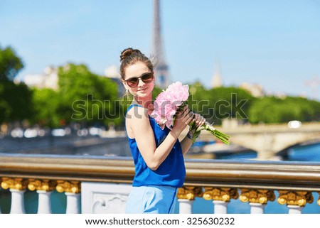 Beautiful young Parisian woman in blue blouse with bunch of pink peonies on the Alexandre III bridge in Paris, Eiffel tower is in the background