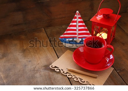 red cup of tea and letter paper next to vintage decorative boat and lantern on wooden old table. retro filtered image
