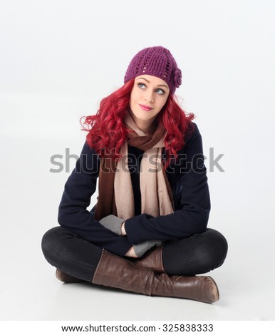 Pretty Red haired girl wearing winter coat, purple woolen hat and scarf,  sitting cross legged. Isolated on white background.
