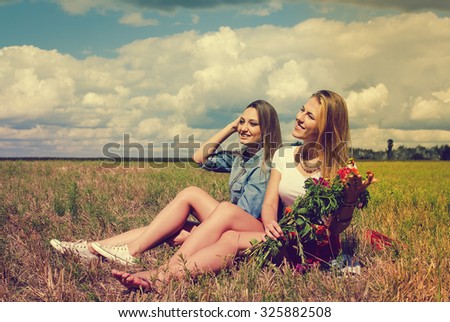 Picture of two beautiful young women sitting on summer meadow. Pretty girls with flower bouquet on retro filtered summer countryside background.