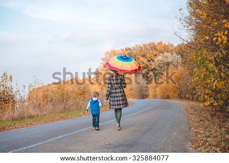 Picture of woman with rainbow umbrella walking with little boy on autumn countryside road. Backview of happy family on golden trees outdoor background.