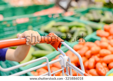 Picture of red handle handcart with boy's hand. Part view of kid taking shopping cart on blurred store vegetable display background.