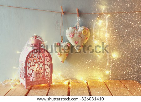 low key image of vintage pink classical frame, fabric hearts hanging on the rope and lantern with garland lights,  on wooden table. retro filtered image with glitter overlay