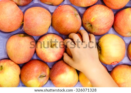 Picture of child's hand taking apple. Store display full of yellow red apples on purple box background.
