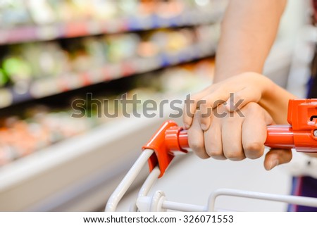 Picture of shopping cart pulling by woman and kid's hands. Part view of happy family beside store display on blurred indoor background.