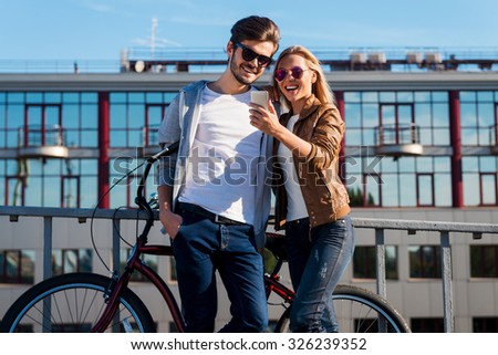 Look at this photo! Beautiful young couple looking at the mobile phone and smiling while standing near bicycle outdoors  Royalty-Free Stock Photo #326239352