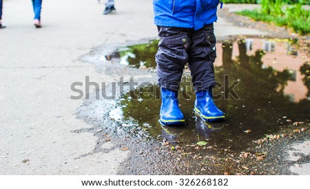 On a rainy day, a boy in blue rubber boots, blue jacket and khaki reel standing in a puddle.