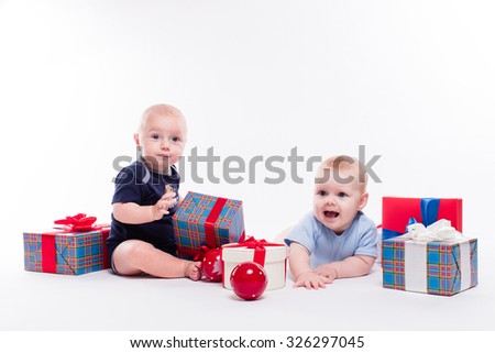 Two cute baby sitting on white background among the Christmas balls and red and blue gifts with smiles on their faces, picture with depth of field