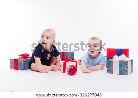 Two cute baby sitting on white background among the Christmas balls and red and blue gifts with smiles on their faces, picture with depth of field
