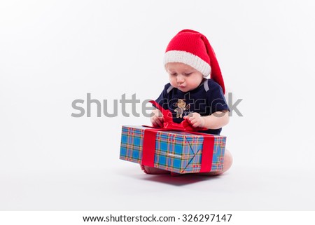 cute toddler is sitting in a New Year's red cap on white background unpacks a gift with a smile on his face, picture with depth of field