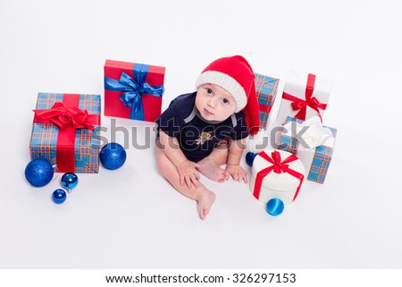 cute toddler is sitting in a New Year's cap among Christmas toys and blue and red boxes with gifts and looking at the camera with a smile on his face, picture with depth of field