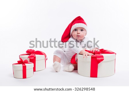 beautiful kid sitting in a New Year's cap and white body among the boxes of Christmas gifts and looking at the camera, picture with depth of field