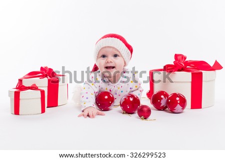 Cute baby lying on his stomach in a New Year's cap among Christmas balls and red box with presents and looking at the camera with a smile on his face, picture with depth of field