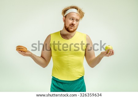 Funny picture of red haired, bearded, plump man on white background. Man wearing sportswear. Man holding apple and sandwich