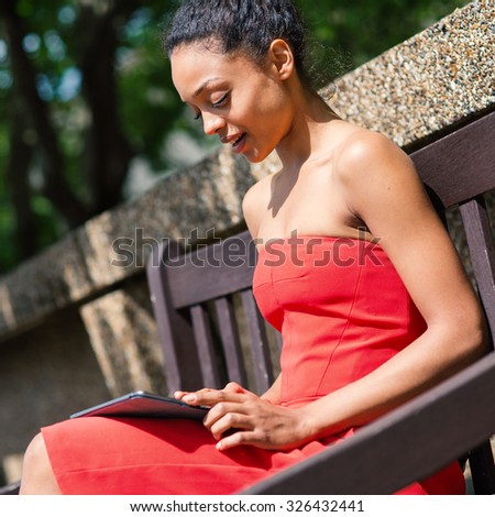 Young mixed race businesswoman portrait outdoors in London while using tablet sit on a bench in a park. Filtered image.