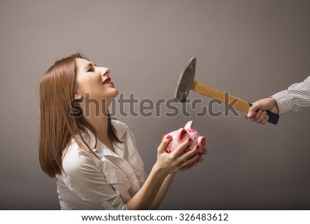 Expressive woman protects her's economy piggy bank of someone who is trying to smash it with a hammer, studio shot on grey background