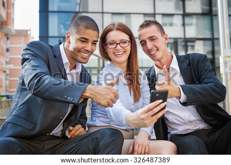 Group of business people sitting at stairs in front of their office and making selife, guys showing thumbs up.