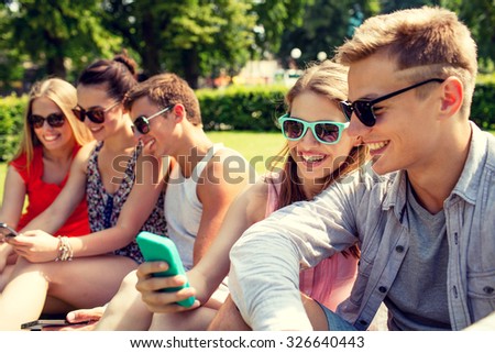 friendship, leisure, summer, technology and people concept - group of smiling friends with smartphone sitting on grass and making selfie in park