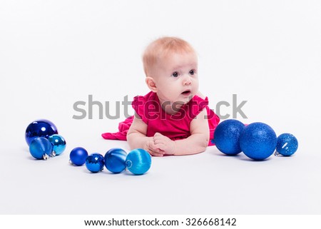 Cute girl lying on her stomach on a white background in a smart red dress among blue Christmas balls, picture with depth of field