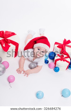 Cute baby lying on his stomach in a New Year's cap among Christmas toys and blue and red boxes with gifts and looking at the camera with a smile on his face, picture with depth of field