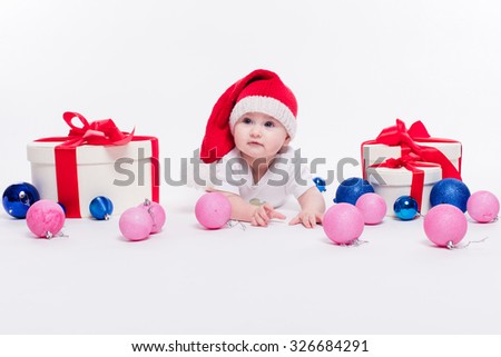 Cute baby lying on his stomach in a New Year's cap among Christmas toys and blue and red boxes with gifts and looking at the camera with a smile on his face, picture with depth of field