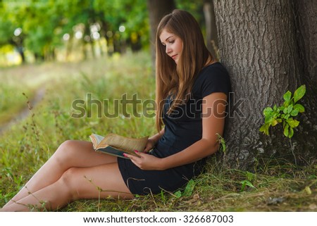 Girl European appearance young brown-haired woman in a black dress with enthusiasm reads the book sitting against a tree, reading, forest