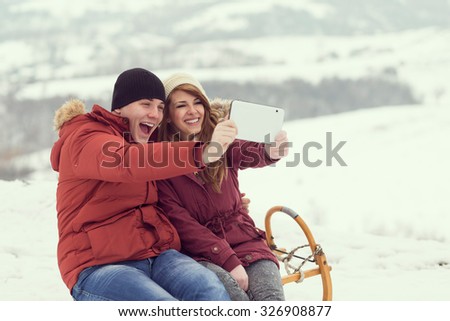 Young couple in love on a winter vacation sitting on a sleigh and making selfie