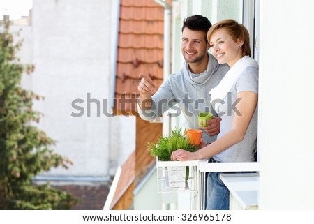 Happy young couple standing at balcony, searching distance, smiling.