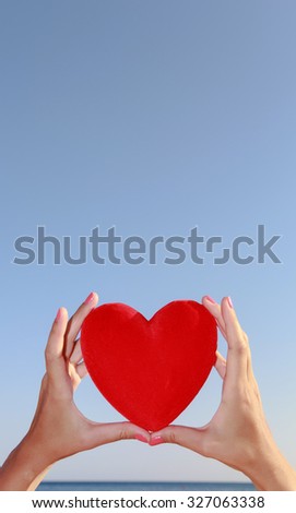 Hands holding red heart with blue sky and sea horizon in the background.